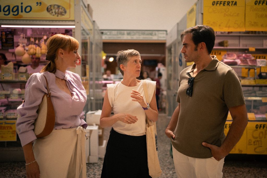 Three people converse in a market, with a woman gesturing as two others listen in Italy