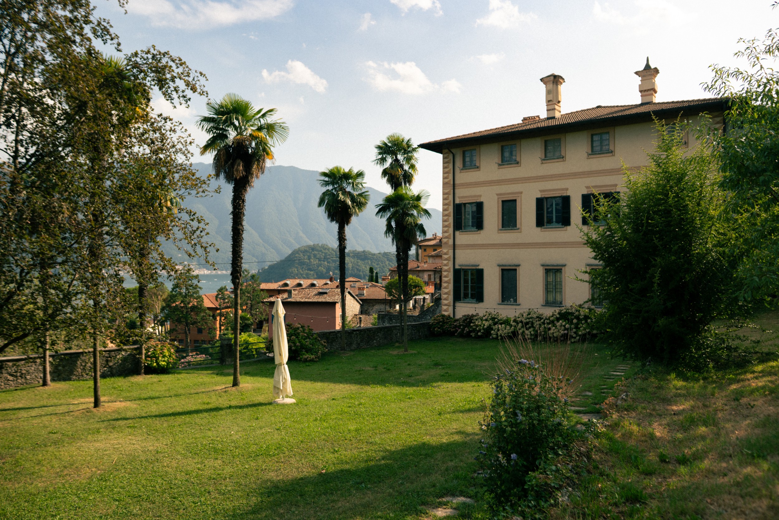 A lakeside Italian village with palm trees, terracotta-roofed houses, mountains, and a large villa