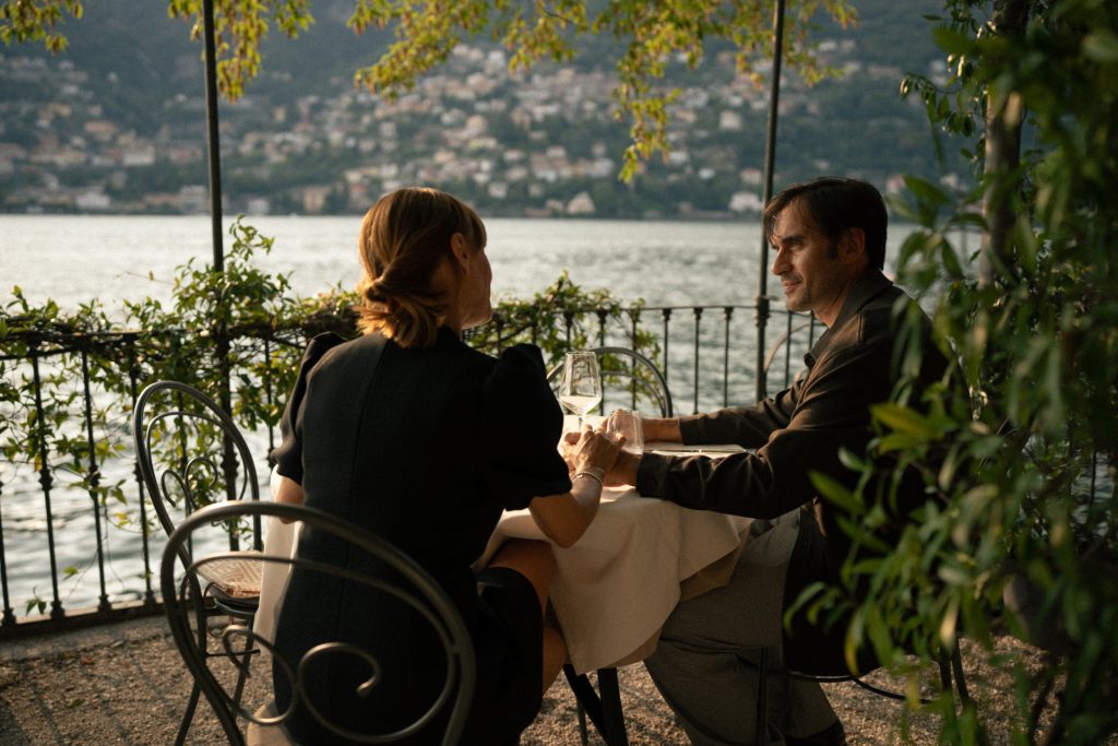 A couple dining at a lakeside restaurant with wine, overlooking a mountain town in Italy