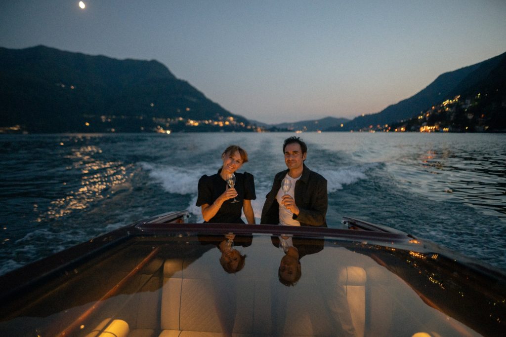 Two people holding champagne glasses on a boat on Lake Como, with a lit mountain lakeside town behind them