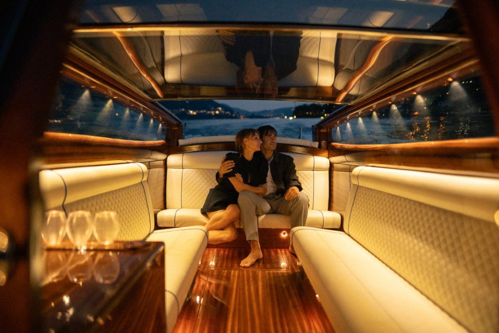 Couple embracing in a warmly lit luxury boat cabin at night, with the water and distant shoreline visible behind them
